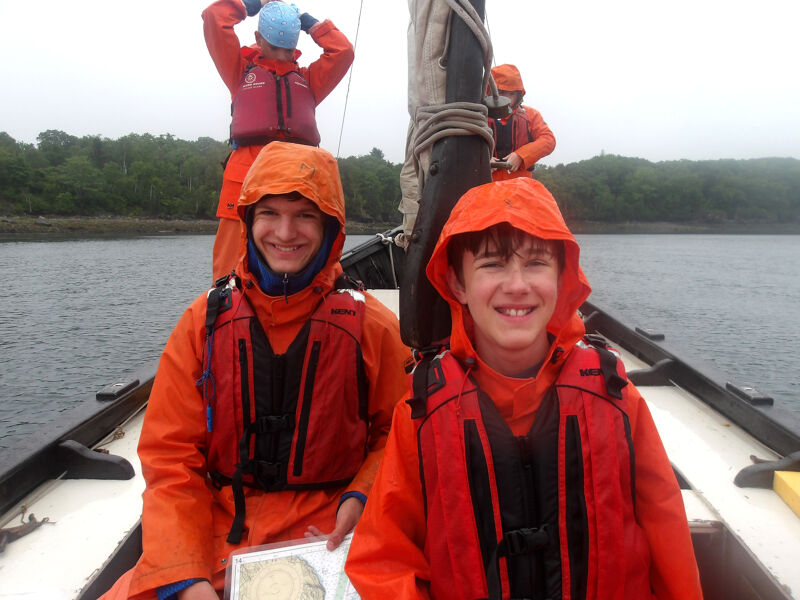 The image shows four people on a boat, likely sailing. Two individuals in the foreground are wearing bright orange hooded jackets and life vests, smiling at the camera. Behind them, another person is adjusting a blue hat, also wearing a life vest and orange jacket. In the background, a fourth person is visible, similarly dressed. The boat is on the water, with trees lining the distant shore, suggesting a river or coastal setting.